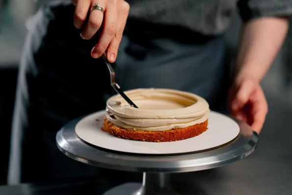 close-up-female-baker-in-a-professional-kitchen-distributes-cream-onto-a-sponge-cake-with-a-spatula-1536x1024