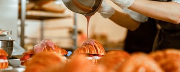 Confectioner pours pink icing onto tasty croissant from metal bowl on counter